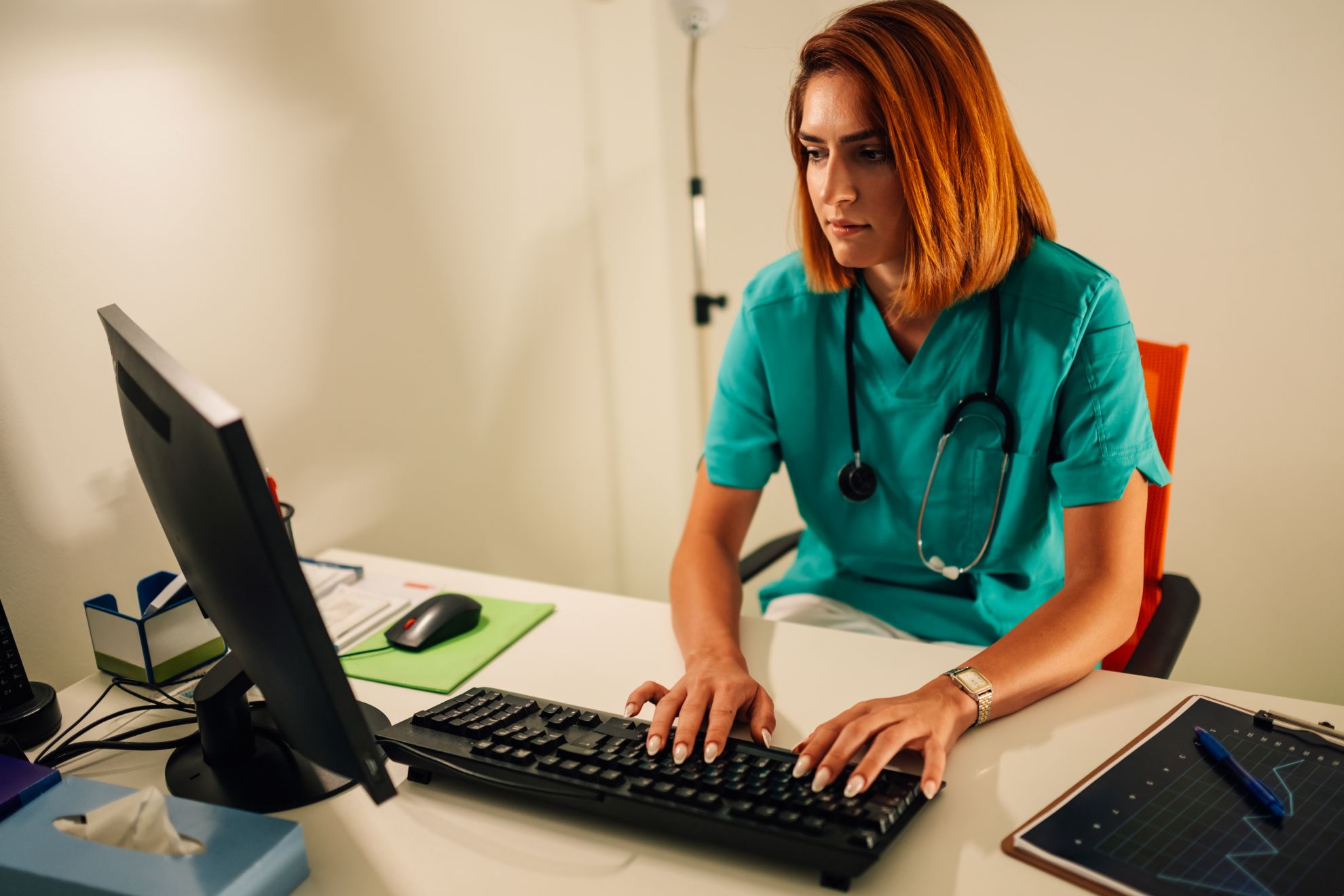Healthcare professional in teal scrubs typing service notes at a desktop computer